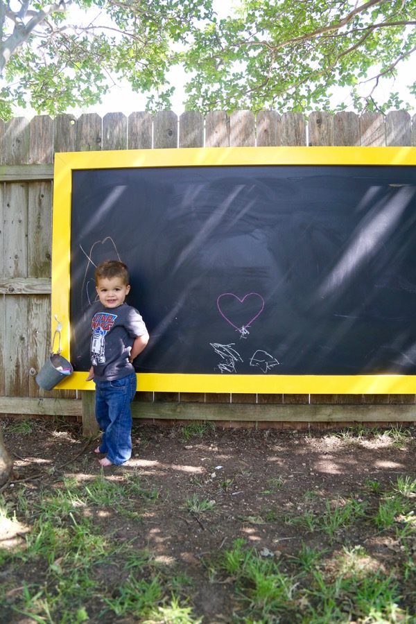 toddler posing in front of an outdoor chalkboard hanging on fence