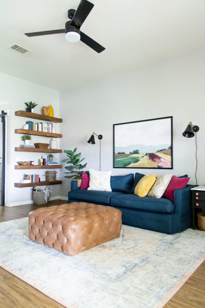 living room with a blue sofa, leather ottoman, and floating open shelving