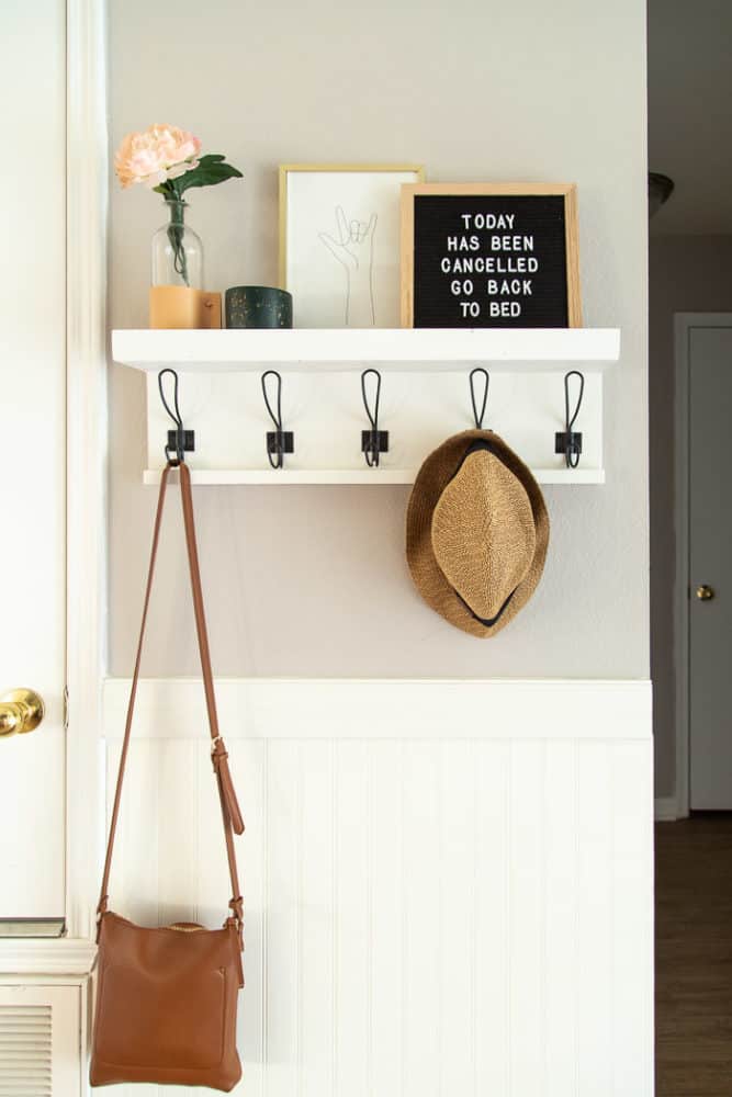 entry to a home with a white wall shelf with hooks, styled with art and a hat hanging on a hook