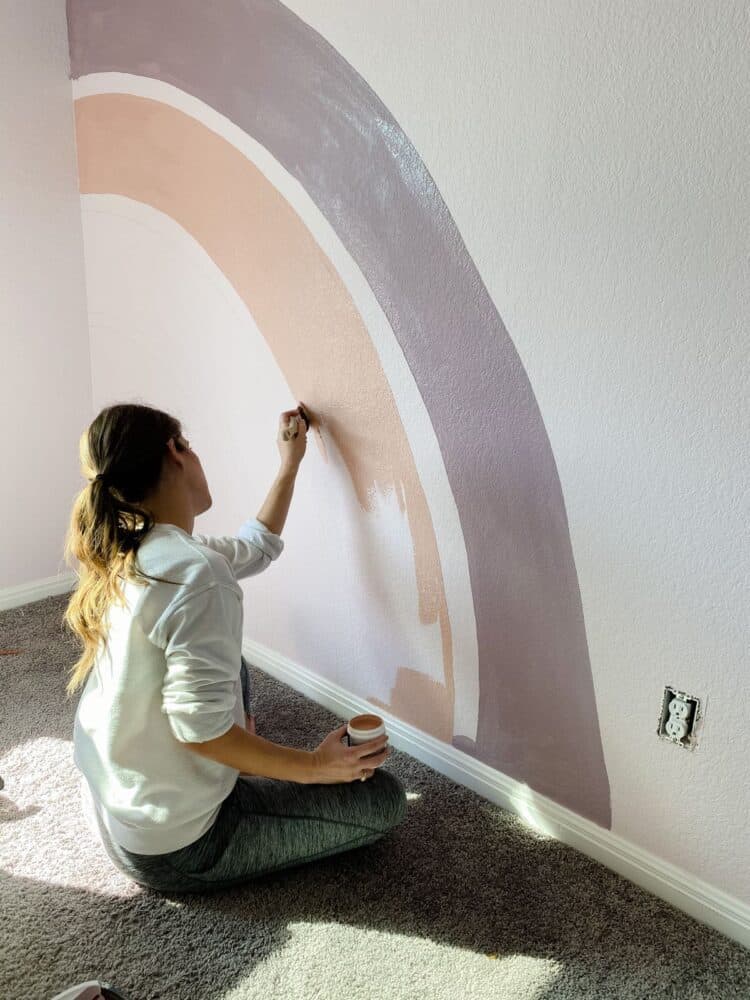 woman painting a rainbow mural on a wall in a little girl's room 