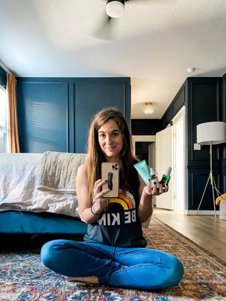 woman sitting in front of a mirror holding items from her skincare routine 