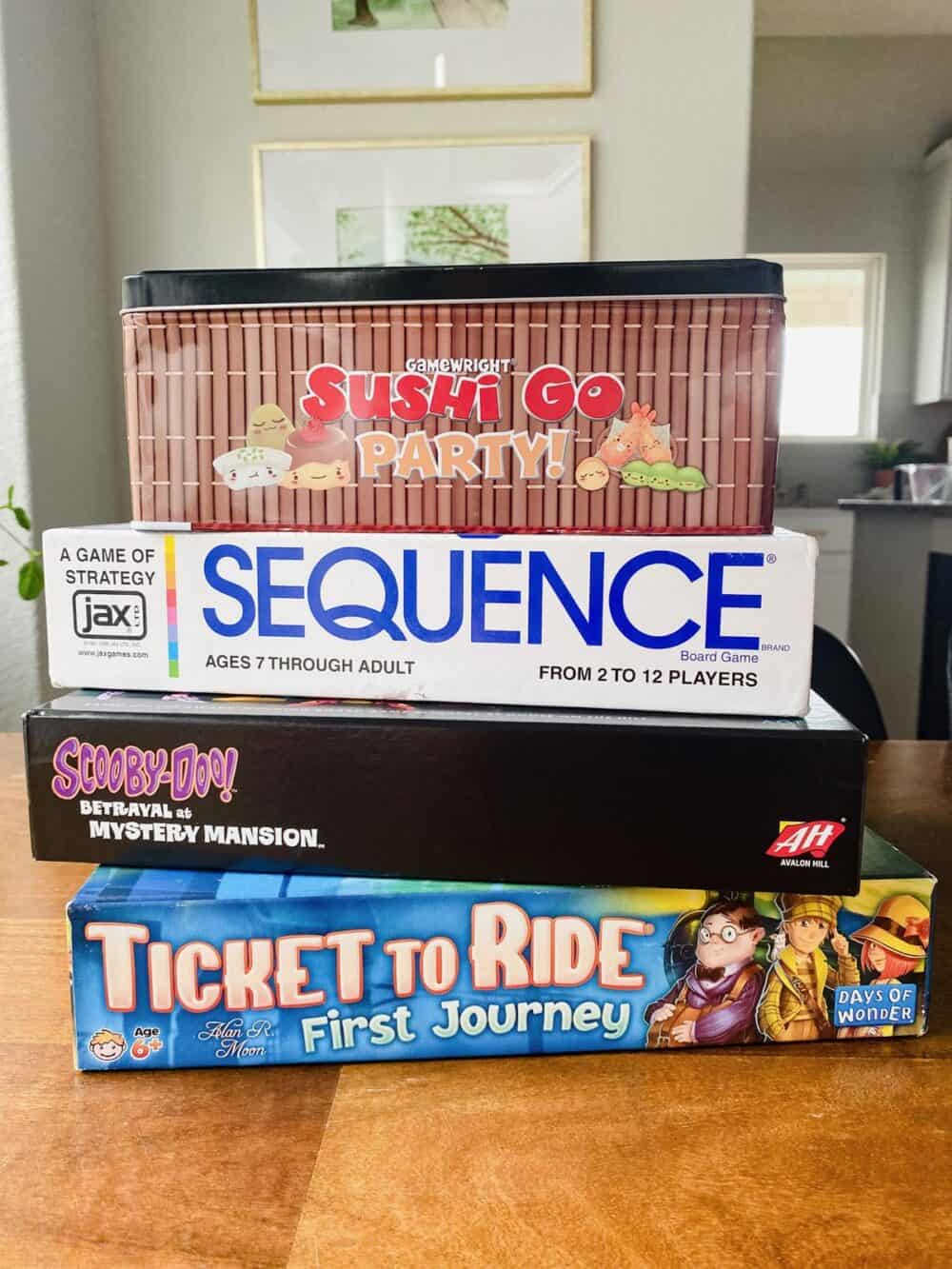 Stack of board games for kids, including Ticket to Ride First Journey, Betrayal at Mystery Mansion, Sequence, and Sushi Go