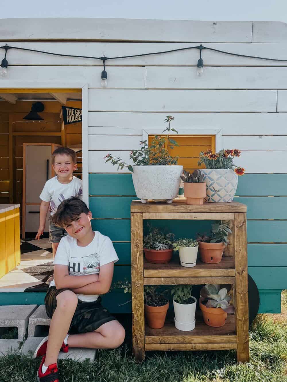 Two young boys in a playhouse camper next to a small plant stand 