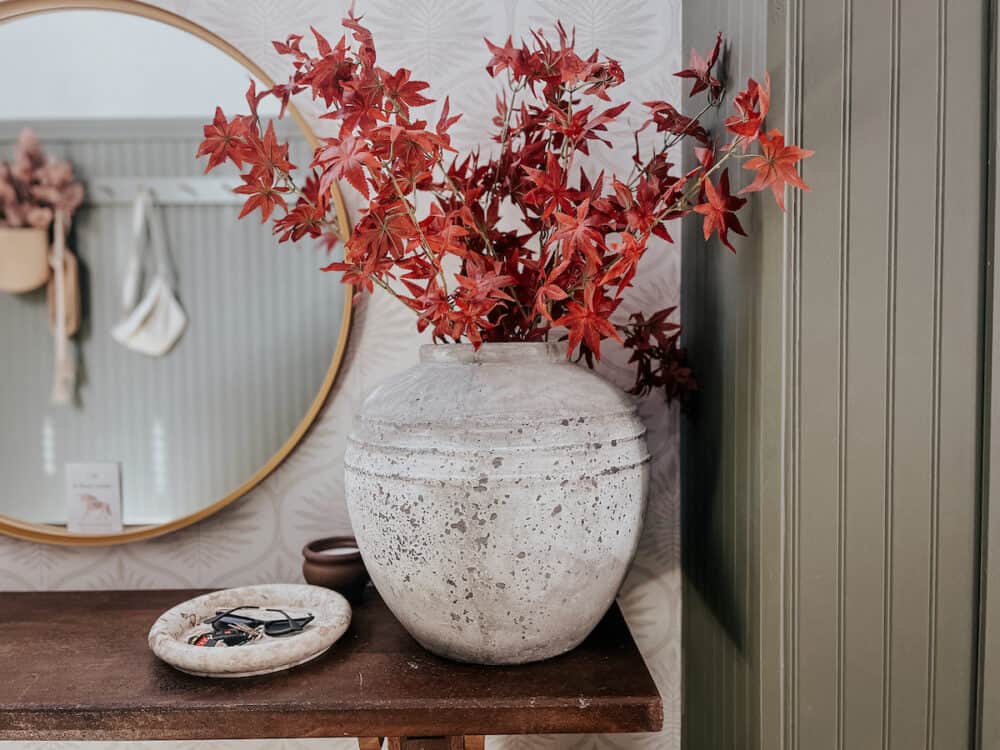 an entry with a large gray vase that has faux maple leaves in it